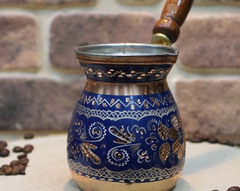 Pode incluir: Uma cafeteira turca de cobre e azul com intrincados desenhos florais. A cafeteira tem uma pega de madeira e está sentada sobre uma superfície de madeira com grãos de café e anis estrelado.