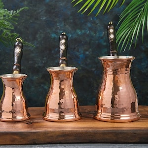 Puede incluir: Tres cafeteras turcas de cobre martillado con mangos de madera. Las cafeteras son de diferentes tamaños y están dispuestas sobre una superficie de madera.