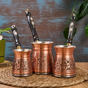 May include: Three copper Turkish coffee pots with ornate designs and black wooden handles. The pots are arranged in a row on a woven placemat.