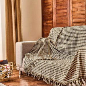 May include: A beige sofa draped with a brown and white checkered throw blanket with fringe. The blanket covers the sofa and hangs over the arm. A gold basket with books and other items sits on the floor.