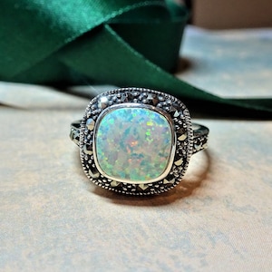 May include: A silver ring featuring a square opal gemstone, displaying iridescent colours. The opal is framed by small, dark stones, with an ornate band design. The ring is set against a neutral background.