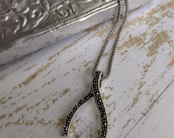 Peut inclure: Un collier argenté avec un pendentif en forme de wishbone orné de petites pierres sombres. La chaîne et le pendentif reposent sur une surface en bois clair vieilli. Une boîte argentée décorative est visible en arrière-plan. Un bijou élégant.