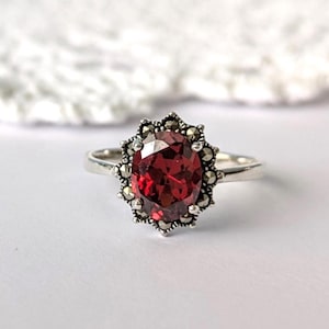 Op de afbeelding: Een zilveren ring met een grote, ovale, dieprode edelsteen. De edelsteen is omgeven door een halo van kleine, ronde, zilverkleurige stenen. De ring is geplaatst tegen een zachte, witte achtergrond.