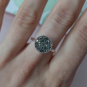 Puede incluir: Un anillo plateado con una cara redonda y oscura, adornada con gemas. El anillo tiene una banda delgada y plateada, y el grupo de gemas está rodeado por un borde con cuentas. El anillo se muestra en un dedo.