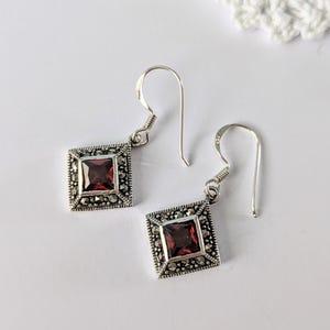 May include: A pair of silver earrings with a square, dark red gemstone center. The earrings have a marcasite border and a hook closure. The design is symmetrical and the earrings are displayed on a white surface.