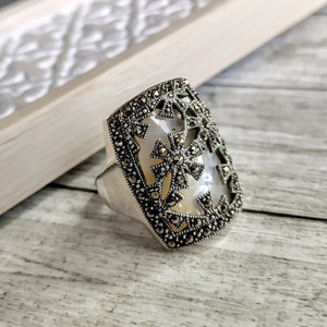 Könnte beinhalten: Silberring mit rechteckiger Oberfläche. Die Oberfläche hat einen Perlmutt-Mittelteil mit dekorativem Markasit-Rand und kreuzartigen Akzenten. Der Ring liegt auf einer hellen Holzoberfläche.