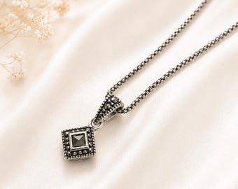 Peut inclure: Un collier en argent avec un pendentif carré orné d'une pierre noire au centre. Le pendentif est entouré de petits détails décoratifs. La chaîne est de conception texturée. Le collier repose sur une surface blanche et soyeuse.