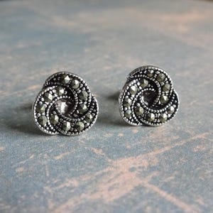 Könnte beinhalten: Ein Paar silberfarbene Ohrstecker. Jeder Ohrstecker hat ein wirbelndes, dreilappiges Design, besetzt mit kleinen, dunklen, facettierten Steinen. Die Ohrringe haben eine Vintage-Ästhetik und eignen sich für den Alltag oder besondere Anlässe. Durchmesser ca. 1 cm.