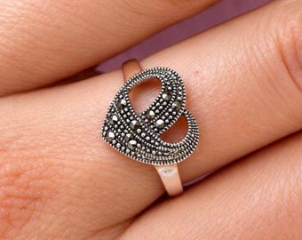 Puede incluir: Un anillo de tono plateado con un diseño en forma de corazón y un centro abierto. La superficie del corazón está densamente incrustada con pequeñas piedras oscuras facetadas que crean una apariencia metálica texturizada. El aro es liso y pulido.
