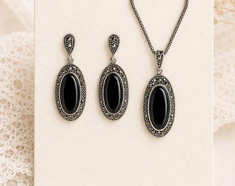 Peut inclure: Un ensemble de bijoux comprenant un collier et des boucles d'oreilles. Chaque pièce présente une pierre noire ovale entourée de petites perles argentées. Le collier a un pendentif assorti, et les boucles d'oreilles sont de type pendantes.