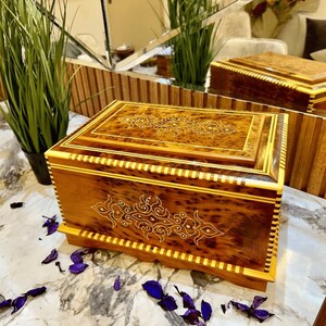 May include: A wooden box with intricate inlaid designs. The box is made of dark wood with light wood accents. The box has a hinged lid and a decorative base.