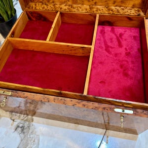 May include: Open wooden jewelry box with a rich, warm wood grain finish. The interior is lined with red velvet and divided into compartments. The box has a rectangular shape with a hinged lid and brass hardware.