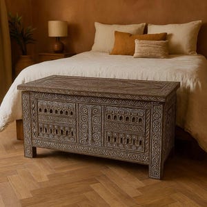 May include: A carved wooden storage chest with intricate white designs. The chest is brown and has a rectangular shape. It is sitting on a wooden floor.