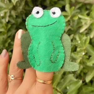 May include: A handmade green felt frog finger puppet with white eyes and black pupils. The frog has a stitched mouth and decorative stitching. The puppet is designed to fit on a finger, and is shown against a background of green foliage.