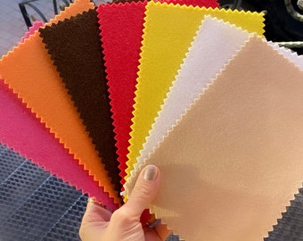 Könnte beinhalten: Eine Sammlung von sieben Filzstoffproben in verschiedenen Farben, darunter Orange, Rosa, Braun, Rot, Gelb, Weiß und Beige. Die Proben sind fächerförmig angeordnet und werden in einer Hand gehalten.