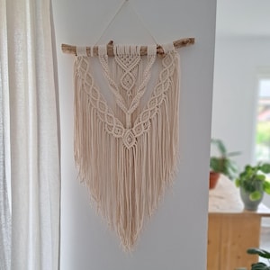 Könnte beinhalten: Beige Makramee-Wandbehang. Das handgefertigte Textilkunstwerk weist komplizierte Knotenmuster und lange Fransen auf, die an einem natürlichen Ast aufgehängt sind. Das Design ist symmetrisch und geometrisch und verleiht der Inneneinrichtung einen Hauch von Bohème.