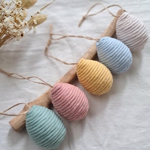 Könnte beinhalten: Fünf handgefertigte Ostereier aus Stoff in Pastellfarben, die an einem Ast hängen. Die Eier sind hellrosa, hellblau, gelb, grün und helllila.