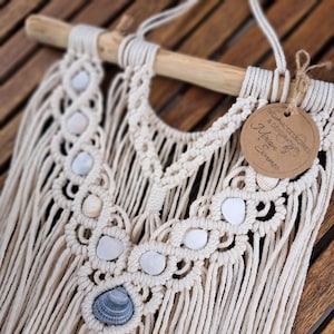 Könnte beinhalten: Cremefarbener Makramee-Wandbehang mit einem Holzdübel. Das Design integriert Muscheln und komplizierte Knotenmuster. Ein braunes Etikett mit Text ist angebracht. Das Stück wird auf einer Holzoberfläche präsentiert.