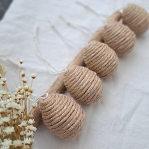 Könnte beinhalten: Fünf natürliche Faserkordel-Ornamente in Tropfenform, die an einem Holzast hängen. Die Ornamente sind hellbraun und haben eine raue Textur.
