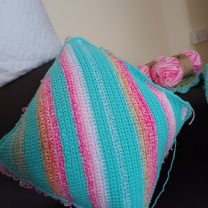 Op de afbeelding: Een gehaakt kussen met een gestreept patroon in turquoise, roze en wit. Het kussen heeft een gehaakte rand.