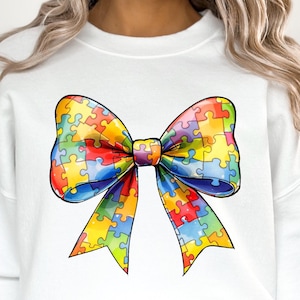 May include: White sweatshirt with a colorful puzzle piece bow design. The bow is made up of puzzle pieces in shades of blue, green, yellow, red, and orange.