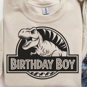 May include: A beige sweatshirt with a black and white graphic of a dinosaur in a Jurassic Park-style logo. The text "Birthday Boy" is printed below the dinosaur.