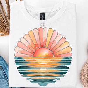 May include: White t-shirt featuring a graphic of a sunset over the ocean. The sunset is depicted with a stylised sun and a series of concentric circles in shades of pink, orange, and yellow. The ocean is represented by a series of blue and green stripes.