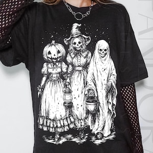 May include: Black graphic t-shirt featuring a Halloween-themed illustration. The design depicts three figures: a pumpkin-headed person, a witch, and a ghost, all with skeletal faces. The artwork is detailed and in a black and white style.