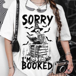 May include: White t-shirt with a black graphic. The graphic shows a skeleton in a witch hat reading a book on a stack of books, with bats and pumpkins. Text reads "SORRY I'M BOOKED".