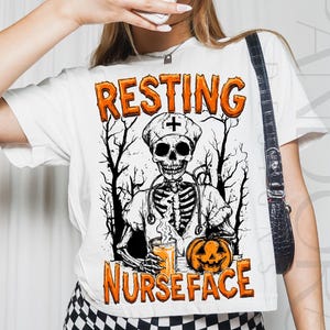 May include: White t-shirt with the words "RESTING NURSEFACE" in orange, featuring a skeleton in a nurse's uniform holding a drink and a jack-o'-lantern. The design includes a medical cross and a forest silhouette, perfect for Halloween.