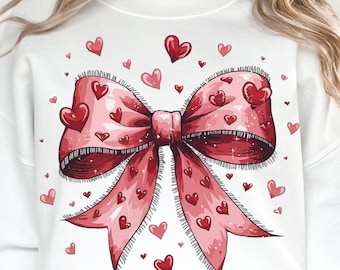 May include: White sweatshirt with a red and pink bow with hearts printed on it. The bow is surrounded by smaller hearts.