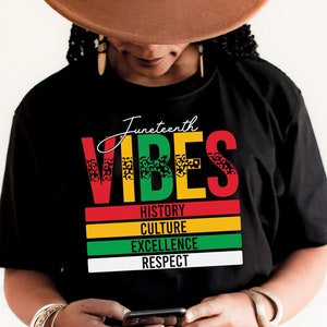 以下が含まれることがあります： レオパード柄のデザインが施されたカラフルなブロックフォントで「Juneteenth Vibes」と書かれた黒いTシャツ。メインテキストの下に「History, Culture, Excellence, Respect」という文字が小さなフォントで印刷されています。