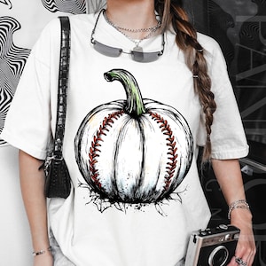 Puede incluir: Camiseta blanca con un diseño gráfico único de una calabaza con forma de pelota de béisbol. La calabaza tiene un tallo verde y costuras rojas, con un diseño artístico en blanco y negro. Una fusión creativa de Halloween y béisbol.