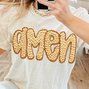 May include: Cream-colored t-shirt with the word "AMEN" in a textured, light brown, crochet-style font. The shirt has short sleeves and a relaxed fit. The text is the focal point, creating a simple yet eye-catching design.