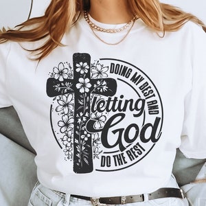 Op de afbeelding: Witte longsleeve met een zwart kruis en een bloemenontwerp. Het shirt heeft de tekst "Doing my best and letting God do the rest" rond het kruis.