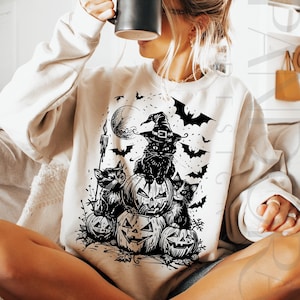 May include: Cream-colored sweatshirt featuring a black and white Halloween design. The graphic includes a black cat wearing a witch hat, perched atop carved pumpkins, with bats, a full moon, and a candle.