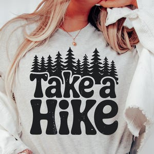 Bold black text saying “Take a Hike” is centered beneath a dense silhouette of pine trees, all in a high-contrast black design on a white background.