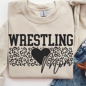 May include: A beige sweatshirt with the text "Wrestling Mom" in black lettering. The text is surrounded by a black leopard print design and a black heart.