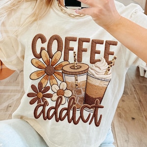 This design features the words 'Coffee Addict' in bold, retro-style lettering in warm brown tones. The graphic features two iced coffee cups complete with straws and whipped cream, surrounded by large floral elements in neutral brown shades. The overall aesthetic is trendy yet cosy, drawing inspiration from boutique-style apparel for coffee lovers.