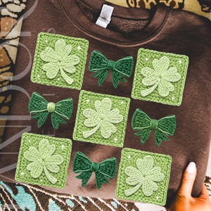 May include: A brown sweatshirt adorned with crocheted green shamrocks and bows. The shamrocks are in square frames, and the bows are in between the shamrocks. The design is a mix of light and dark green. The sweatshirt is held by a person.