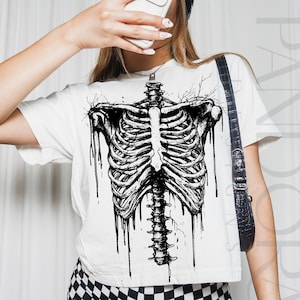 Może przedstawiać: Biała koszulka z czarno-białą ilustracją ludzkiej klatki piersiowej i kręgosłupa. Grafika ma efekt kapania. Modelka ma na sobie czarny kapelusz i spodnie w kratę.