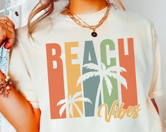 May include: A light beige t-shirt with a retro-style graphic design featuring the text "Beach Vibes" and palm trees in shades of orange, yellow, green, and white.