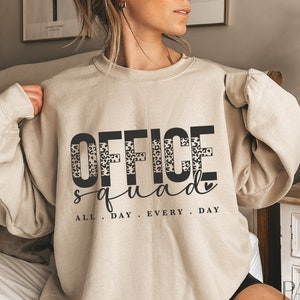 Peut inclure: Un sweat-shirt beige avec le texte "OFFICE squad ALL. DAY. EVERY. DAY" imprimé sur le devant. Le mot "OFFICE" est imprimé en noir avec un motif léopard derrière les lettres.