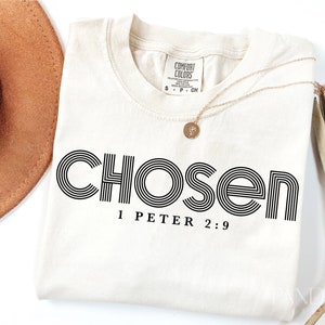 Puede incluir: Una camiseta de color crema con texto negro que dice "chosen 1 Peter 2:9". El texto está en una fuente retro y en negrita.