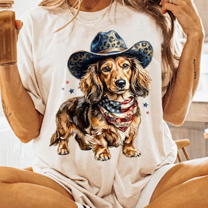 May include: A beige t-shirt featuring a dachshund wearing a cowboy hat with a leopard print and a bandana with an American flag design. The dog is brown and tan with long fur. Red and blue stars are scattered around the dog.
