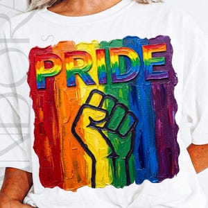 May include: White t-shirt featuring a colorful Pride design. The design includes the word "PRIDE" in rainbow colors, a raised fist, and a rainbow-colored background. The artwork has a textured, painted appearance.