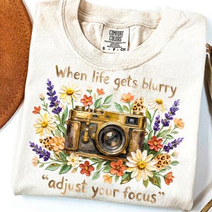 May include: Cream-colored t-shirt with a floral and camera design. The graphic features a vintage camera surrounded by colorful flowers and the text "When life gets blurry, adjust your focus."