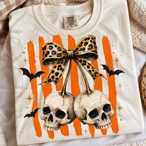 Two skulls hang like cherries under a leopard print bow, surrounded by orange brushstroke stripes, black bats, gold stars, and textured Halloween details.