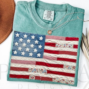 May include: A sea green t-shirt with a sewn-on American flag design. The flag features denim blue with white stars and red and floral print stripes. The shirt has a Comfort Colors tag and a silver necklace with a pendant.