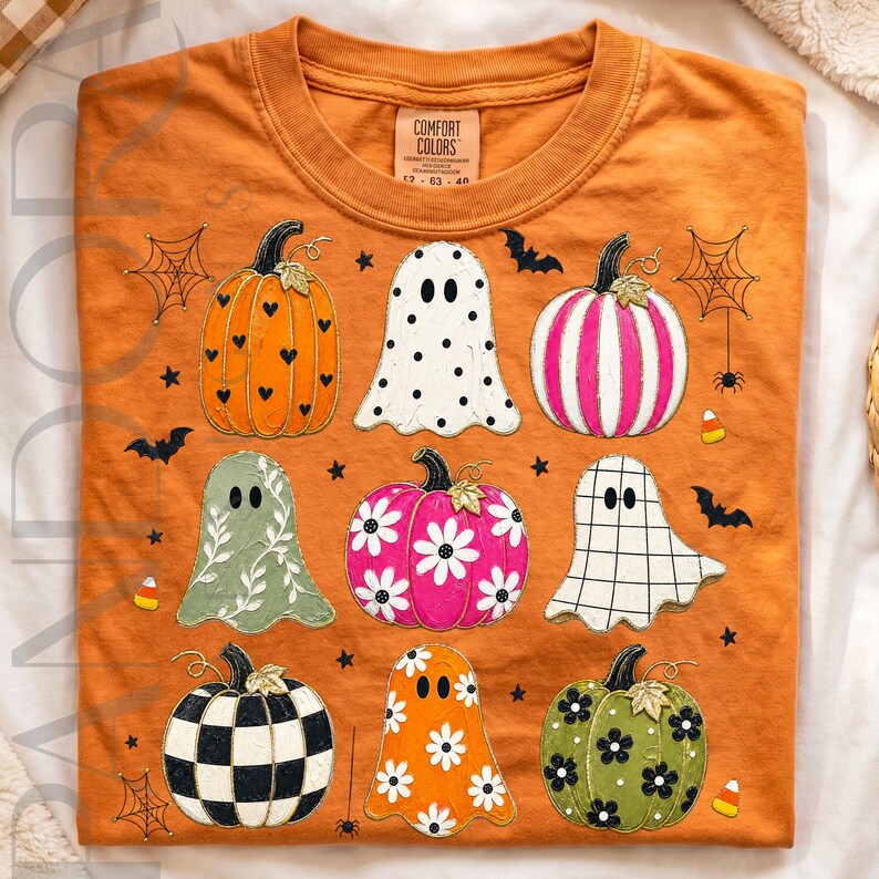 Op de afbeelding: Een oranje t-shirt met een grafische print van diverse pompoenen en spoken. De pompoenen zijn oranje met zwarte hartjes, roze met witte strepen, roze met witte madeliefjes, zwart-wit geblokt en groen met zwarte bloemen. De spoken hebben stippen, witte ranken en een ruitpatroon.